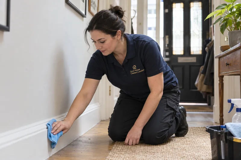 A professional cleaner kneeling comfortably to wipe a painted skirting board — cleaning work in Ilford (IG1 / IG2).